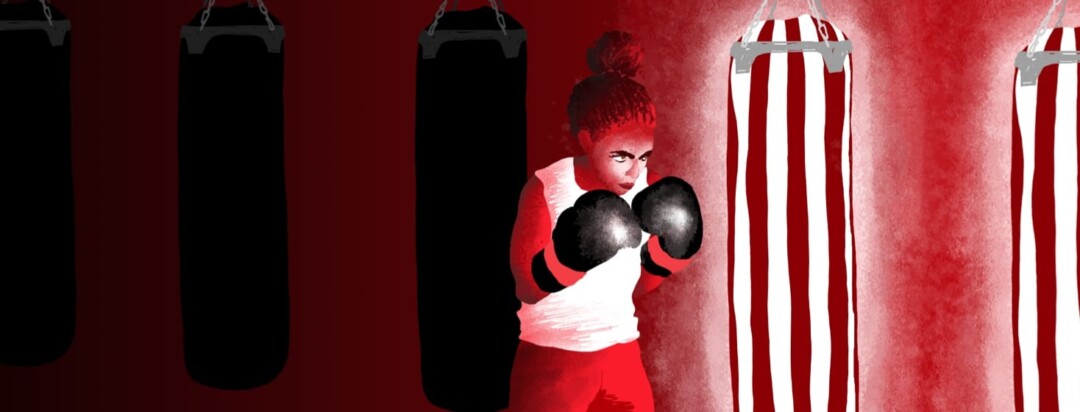 A woman in boxing gloves looking determined at a highlighted pair of punching bags.